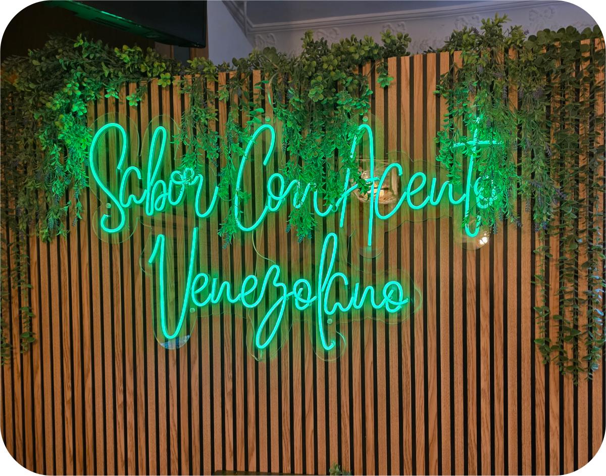 Neón verde Sabor Con Acento Venezolano sobre pared de listones y vegetación